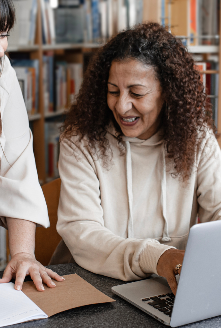 To kvinder står ved et bord i et bibliotek, den ene skriver i en notesbog, mens den anden arbejder på en bærbar computer – begge smiler og ser engagerede ud.