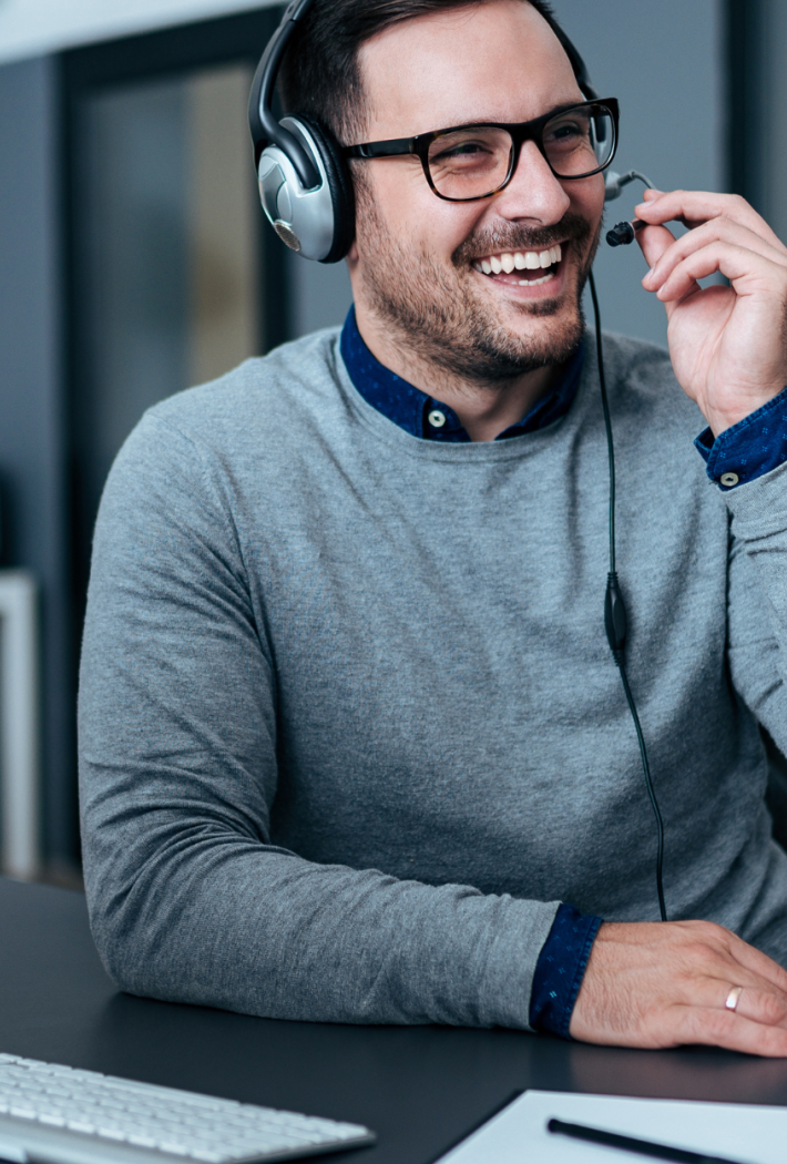 Mand med headset sidder ved et skrivebord og smiler under en samtale foran en stor computerskærm, med bøger og planter i baggrunden.