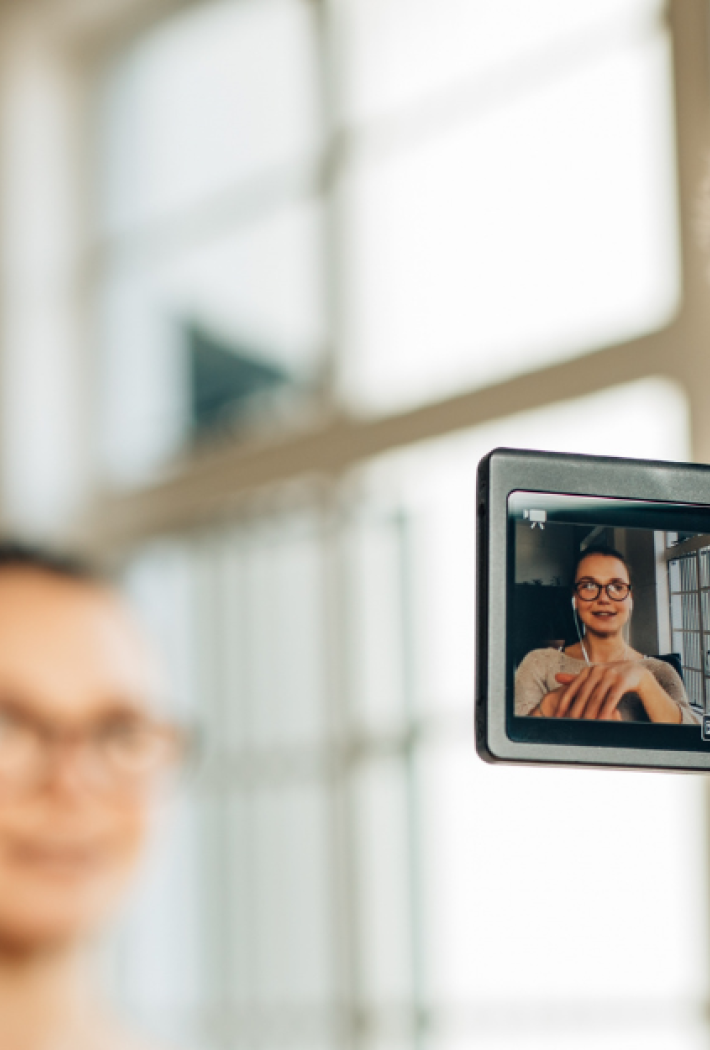 Et kamera på stativ filmer en person med briller og høretelefoner, der vises tydeligt på kameraskærmen. Personen sidder foran et stort vindue og taler til kameraet.
