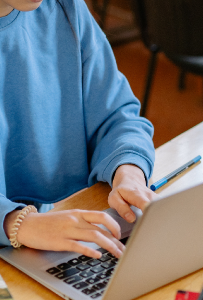 En person sidder ved et bord på et bibliotek og arbejder på en bærbar computer. På bordet ligger åbne bøger, andre studiematerialer og en kuglepen.