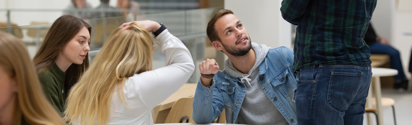 Billedet viser en gruppe studerende i en lys studiehal; en kvinde og en mand sidder ved et bord en stående mand; en bibliotekar,  taler med de studerende. Andre personer studerer i baggrunden.