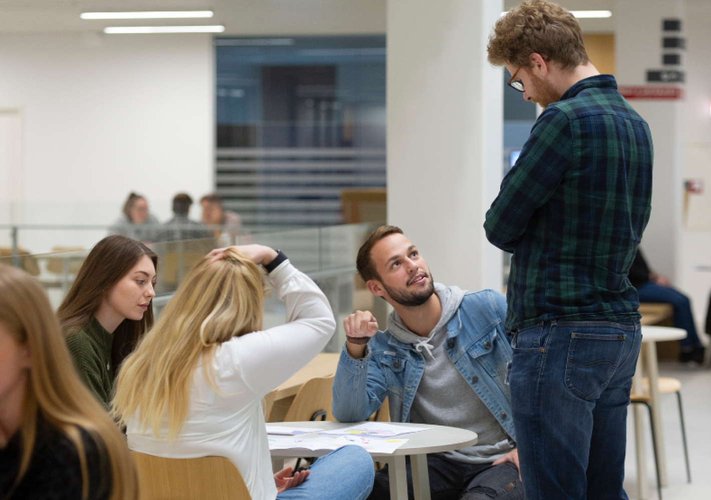 Billedet viser en gruppe studerende i en lys studiehal; en kvinde og en mand sidder ved et bord en stående mand; en bibliotekar,  taler med de studerende. Andre personer studerer i baggrunden.