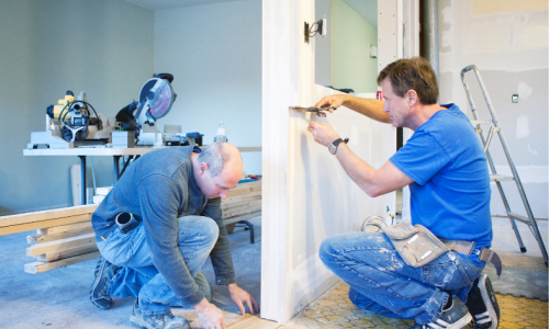 Billedet viser to mænd arbejder i et rum under renovering: den ene skærer træ med en bordrundsav til venstre, mens den anden maler en dørkarm til højre.